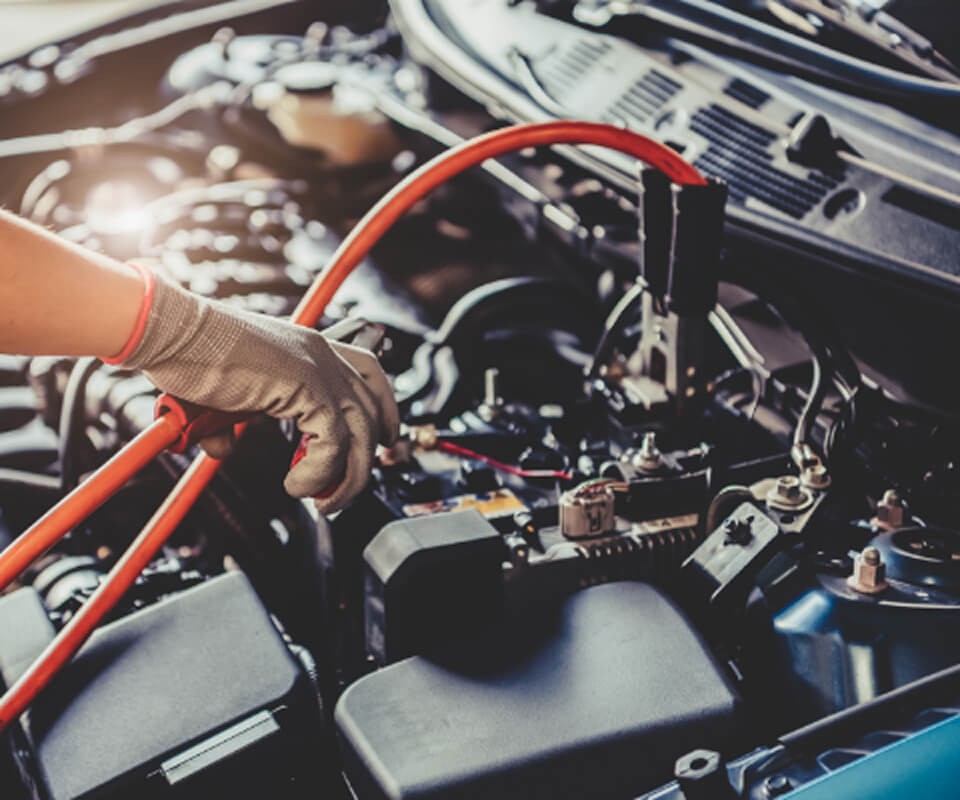 車のバッテリー充電作業の場面。作業者が安全にジャンパーケーブルを接続している様子で、エンジンルーム内の複雑な配線や部品が背景に見える