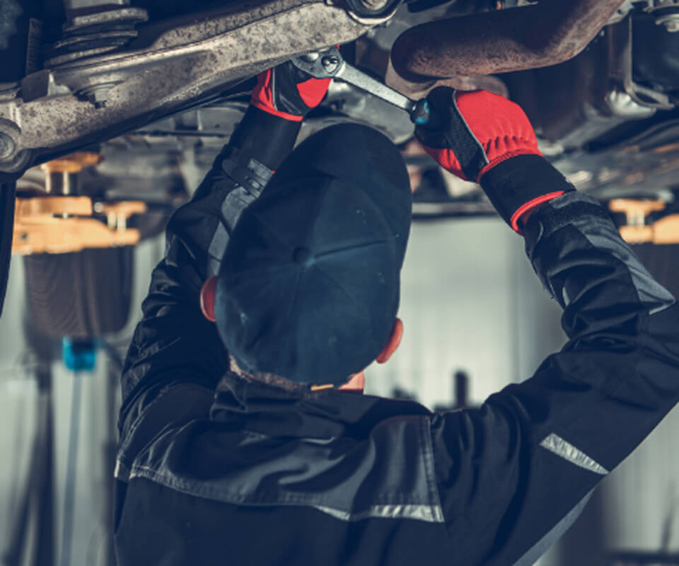 車の下側での整備作業。整備士が専門工具を使って車の部品を修理している様子で、自動車整備の専門的な作業環境