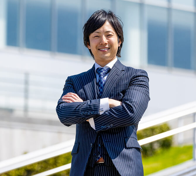 腕を組んで立つ男性のビジネス写真。ピンストライプスーツを着て、明るい笑顔でカメラを見つめている。